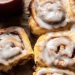 Freshly baked apple cinnamon rolls with a generous layer of white icing, accompanied by cinnamon sticks on a wooden surface.