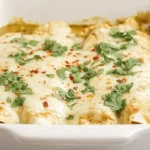 Close-up of bubbling baked green chili chicken enchiladas topped with melted cheese, fresh cilantro, and red pepper flakes in a white ceramic dish.